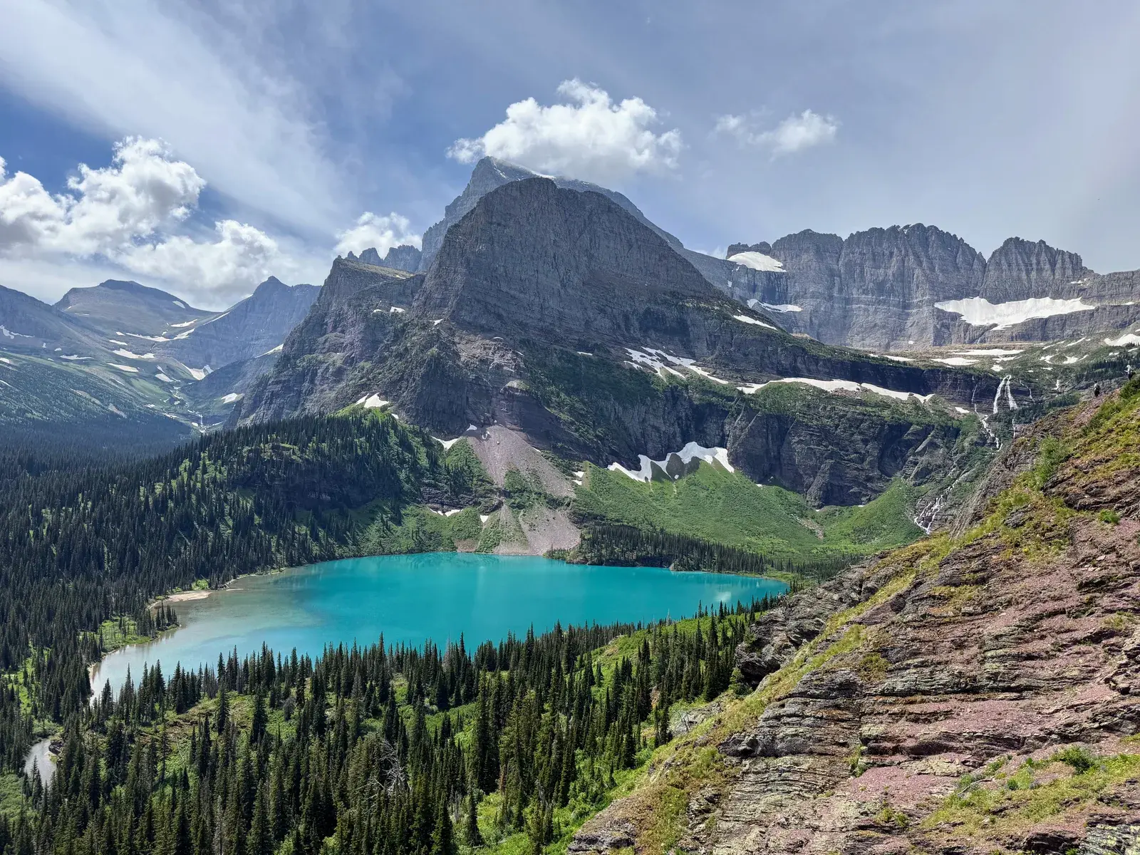A turquoise lake surrounded by pine forests, rugged peaks, and snowfields in Glacier National Park